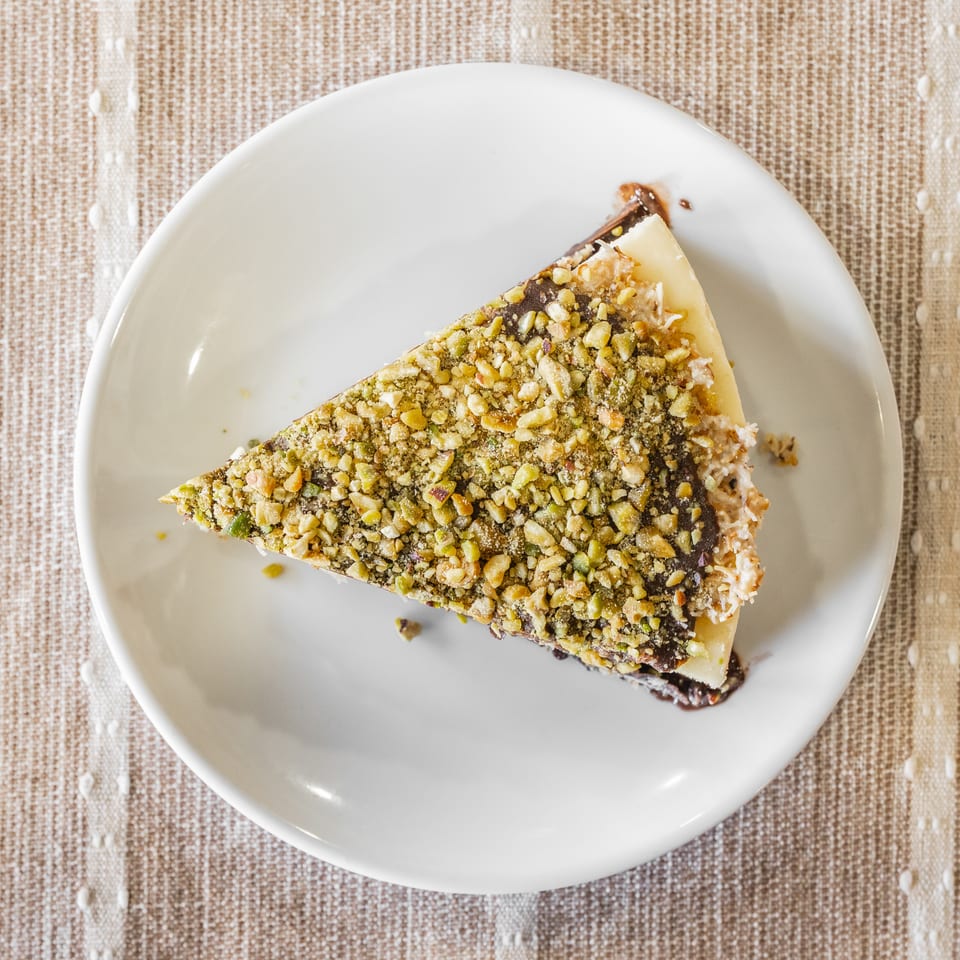 Slice of nut-covered layered dessert on a white plate atop a woven beige tablecloth.