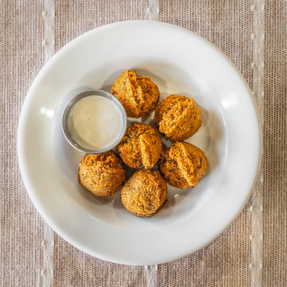 Seven golden-brown falafel balls and a small metal cup of white dipping sauce on a white plate.