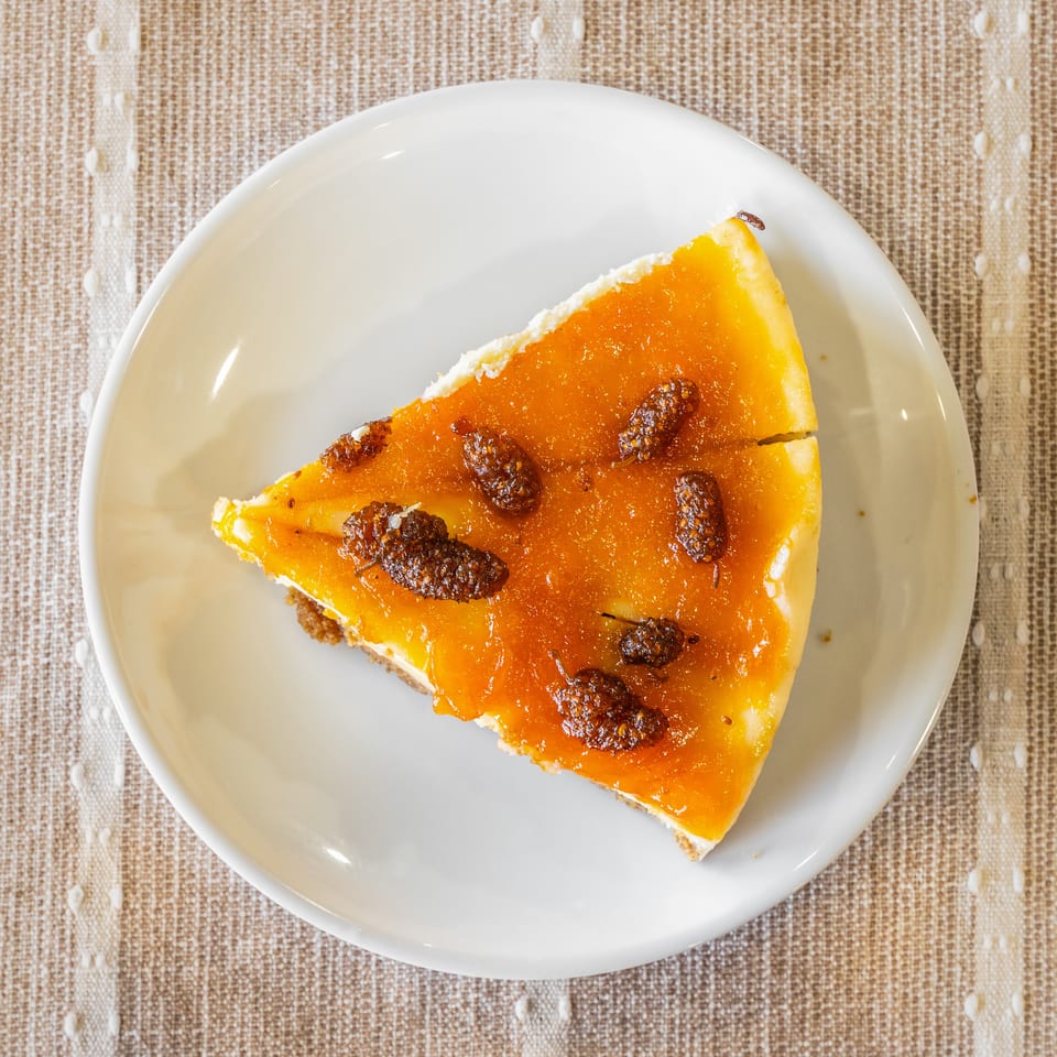 Overhead view of a slice of cheesecake with orange topping and dried mulberries on a white plate.